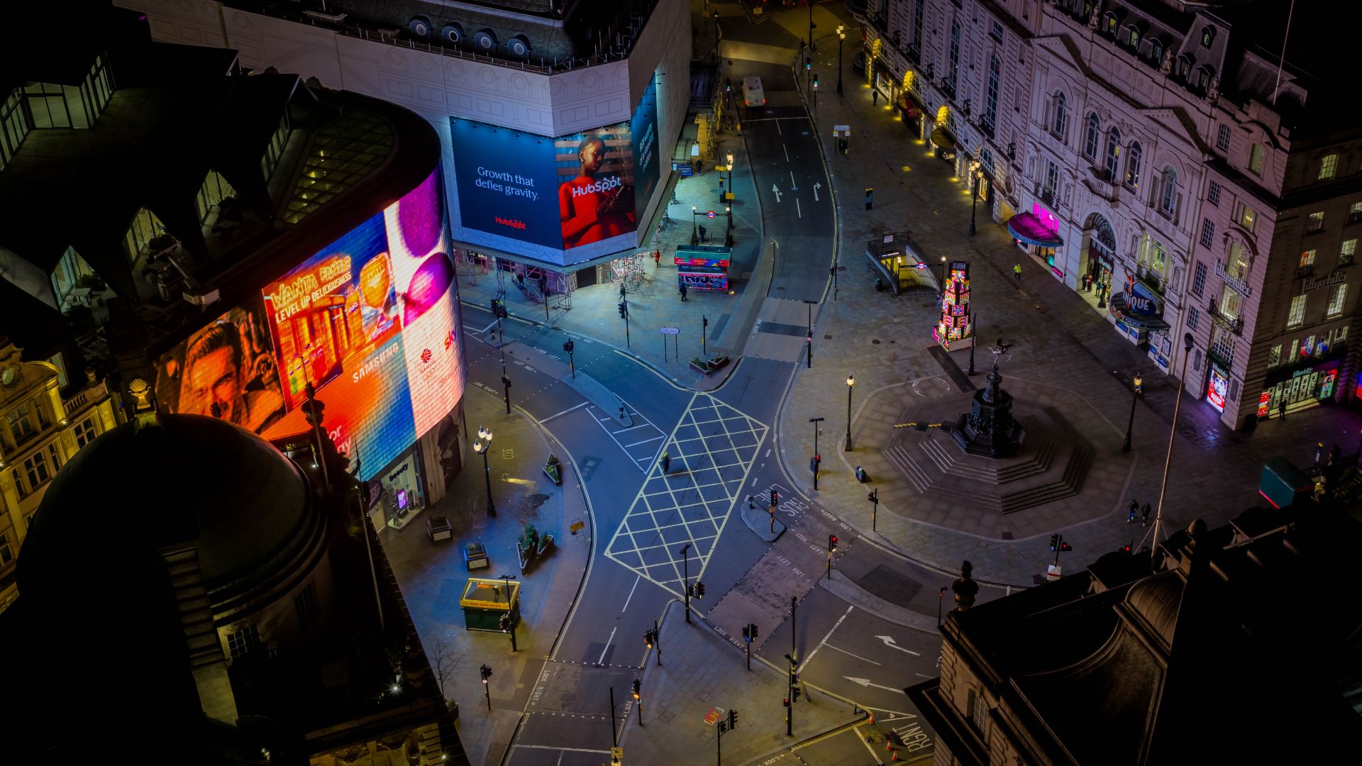 Piccadilly Circus illuminated at night during Art After Dark, with large-scale digital artworks lighting up the famous screens and the usually busy junction momentarily quiet. Image courtesy of Premier Comms. Aerial night view of Piccadilly Circus in London, showing illuminated digital advertising screens, colourful art displays, empty streets, and surrounding historic buildings.