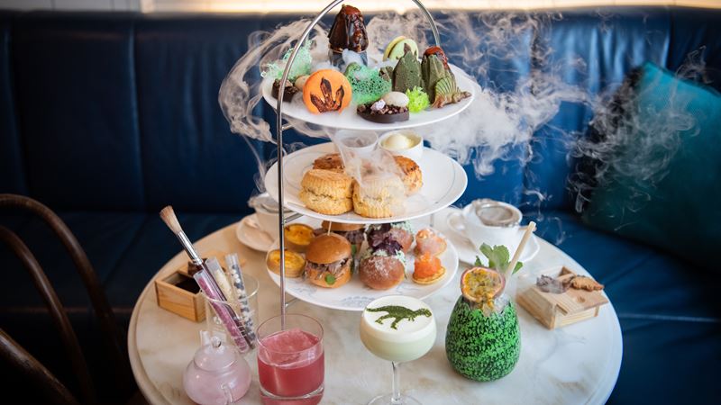 Enjoy a fresh take on tradition with plenty of fun themed afternoon teas in London. Image courtesy of Red Letter Days. Jurassic-themed afternoon tea treats, scones, bites and drinks with smoke and blue seating in the background.
