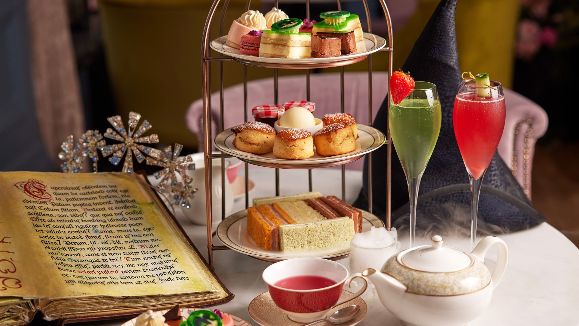 A themed Wicked afternoon tea served at The Clermont London, Victoria, featuring colourful cakes, sandwiches and theatrical cocktails inspired by the musical. Image courtesy of The Clermont Hotel. A tiered afternoon tea stand with cakes, scones and sandwiches beside colourful cocktails and a teapot on a decorated table.