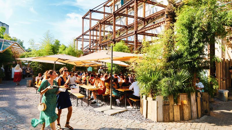 Absorb the rays of sunshine at the open-air Mercato Metropolitano, home to German KRAFT Brewery. Credit: Indira Bott. Image courtesy of German KRAFT Brewery. A photo of two female friends wearing dresses walking along a path on a bright sunny day at Mercato Metropolitan with people sat on picnic benches and parasols and plants in frame