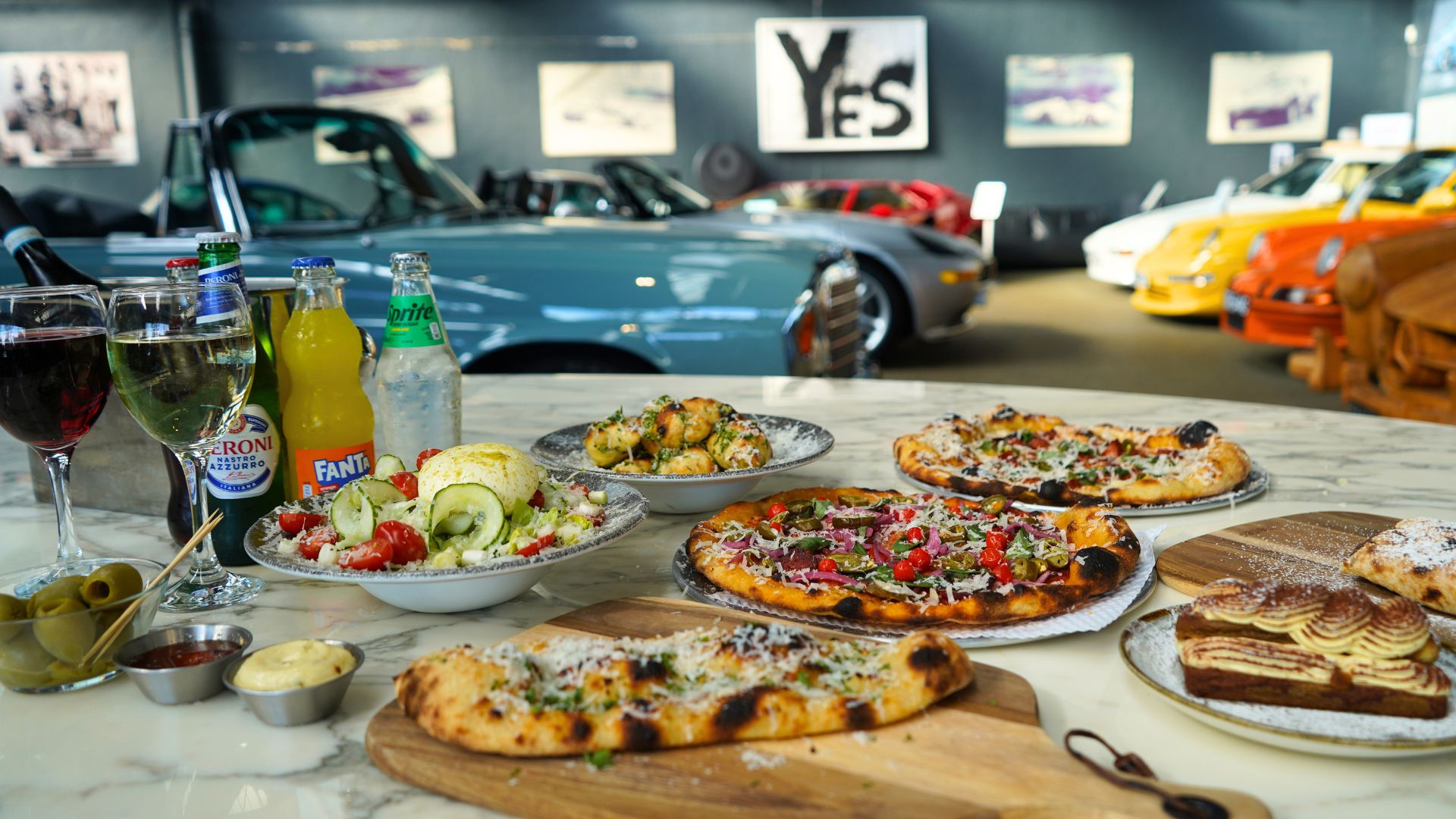 Turn dinner into an experience at 67 Sourdough, where artisan pizzas and indulgent plates are served alongside a backdrop of classic cars – making it just as exciting for little ones as it is for grown-ups. Image courtesy of 67 Sourdough. A table filled with pizzas, salads, drinks and desserts set in front of a display of vintage cars inside a restaurant.