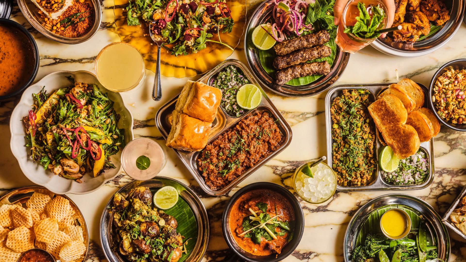 A spread of Bombay-inspired dishes at Dishoom, known for its lively atmosphere and flavour-packed Indian small plates perfect for sharing. Image courtesy of Dishoom. Multiple Indian dishes including curries, breads, kebabs and salads arranged on plates across a marble table.