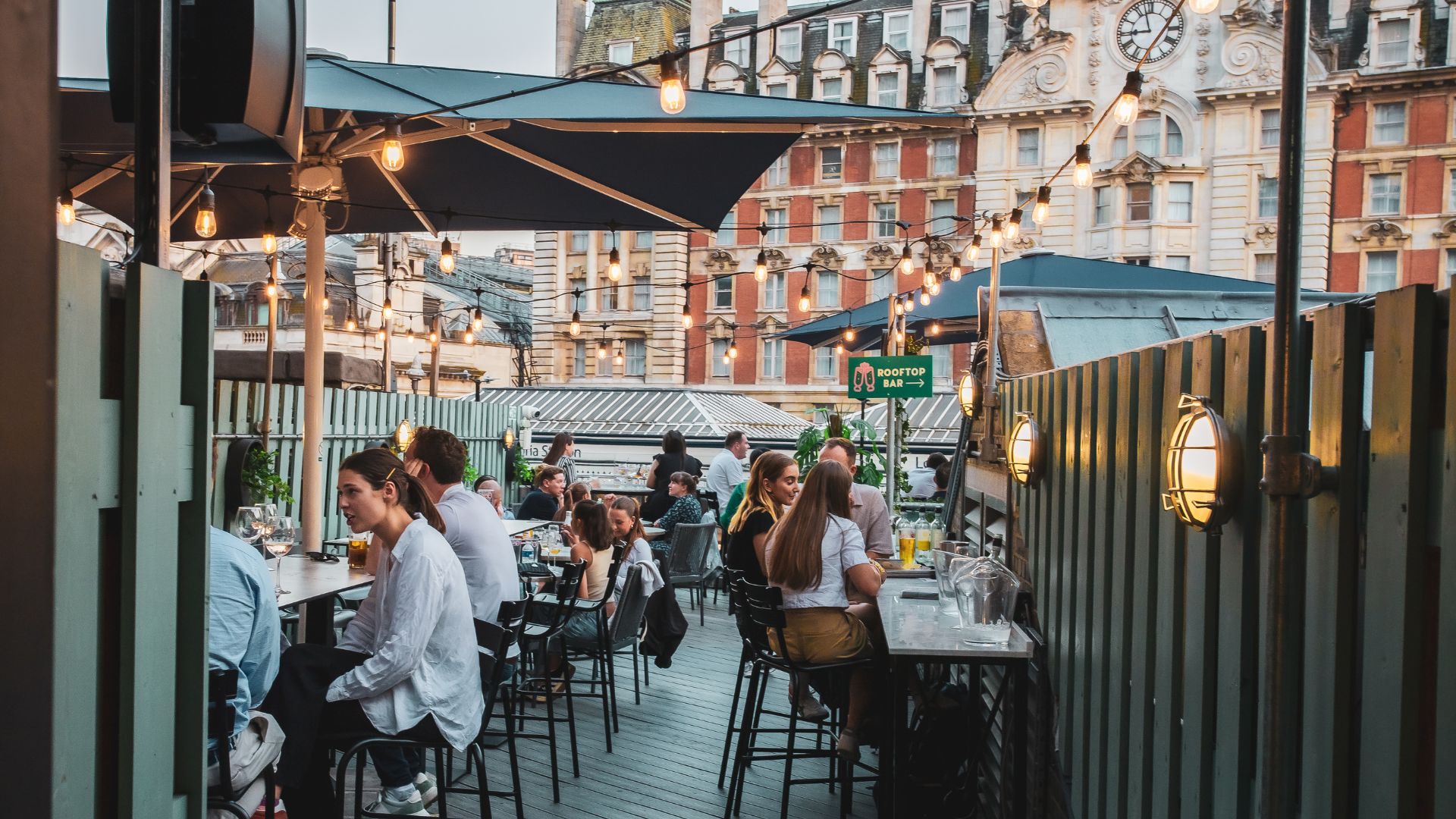 Lively food halls such as Market Halls bring the buzz of London dining to families, with rooftop terraces, long tables and a choice of street-food favourites for every taste. Image courtesy of Market Halls. People sitting at tables on a rooftop terrace with string lights and umbrellas at a busy food hall.