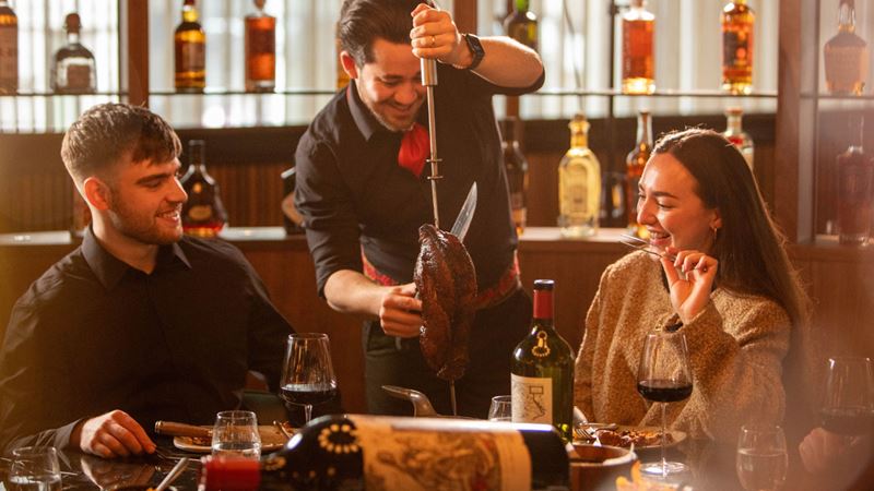 Let the food keep flowing at Fogo de Chão with its menu of prime meats and mouthwatering sides. Credit: Fogo de Chão. Image courtesy of Fogo de Chão. A photo of a couple sat at a table with a glass of wine each and a waiter carving a joint of meat at the table and serving for them
