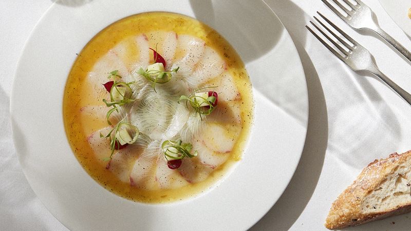 Tuck into succulent Mediterranean-inspired dishes at La Petite Maison in Mayfair. Credit: La Petite Maison. Image courtesy of La Petite Maison. Fish carpaccio drenched in olive oil with slice of bread and forks in the background.