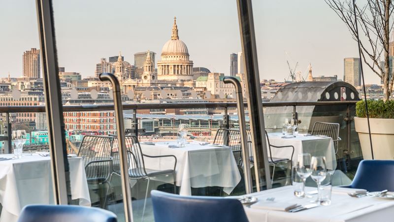 Dine with an incredible view of St Paul's Cathedral at OXO. Credit: OXO Tower Restaurant, Bar and Brasserie. Image courtesy of OXO Tower Restaurant, Bar and Brasserie. Restaurant tables with white tablecloth, wine glasses and blue chairs overlooking the London skyline with St Paul's Cathedral in the background.