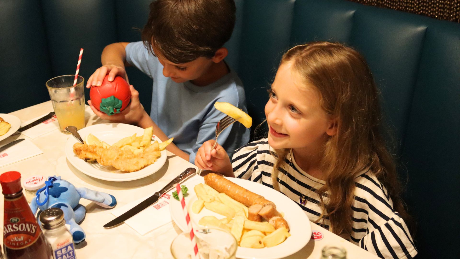 Children enjoy classic fish and chips at Poppies Fish and Chips, where family-friendly portions and retro décor make it a fun spot for younger diners. Image courtesy of Poppies. Two children sitting in a restaurant booth eating fish and chips and sausages with drinks and condiments on the table.