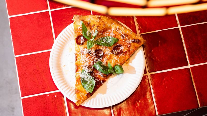 Enjoy a slice or three at Bad Boy Pizza Society spots across London. Credit: Josh Oliver/Bad Boy Pizza Society. Image courtesy of Bad Boy Pizza Society. A bird's eye photo of a slice of pizza on a white paper plate on red tiles, covered in vodka sauce, pepperoni and basil leaves with a sprinkle of fresh parmesan