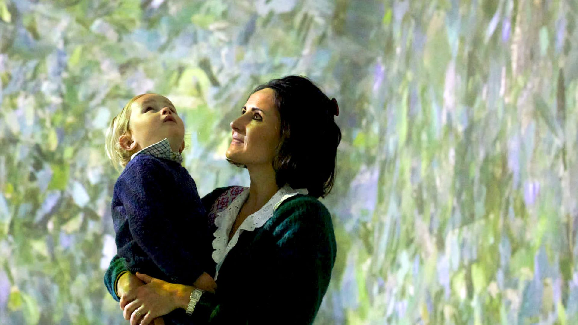 Toddlers will be captivated by the installations at FRAMELESS. Image courtesy of FRAMELESS A woman holding a small child looking up at a leafy green light installation at FRAMELESS in London.