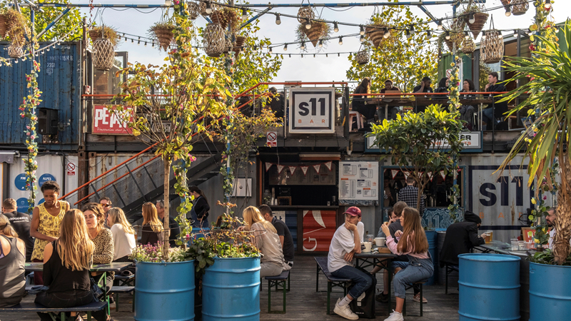 Make memories with friends and family at Pop Brixton, boasting a wide range of food options. © visitlondon.com/Gary Latham. Image courtesy of visitlondon.com People sat outside on benches at Pop Brixton market surrounded by greenery