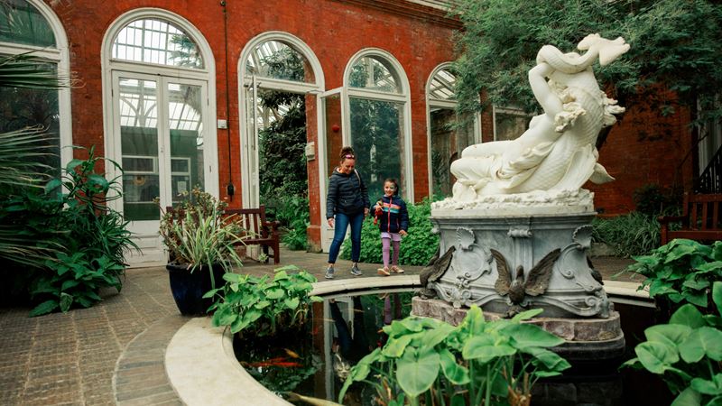 Explore the grounds of Eltham Palace and Gardens year-round. Credit: Visit Greenwich. Image courtesy of Visit Greenwich. A photo of a woman with her young child walking around the winter garden at Eltham Palace with a large water feature in the forgeround