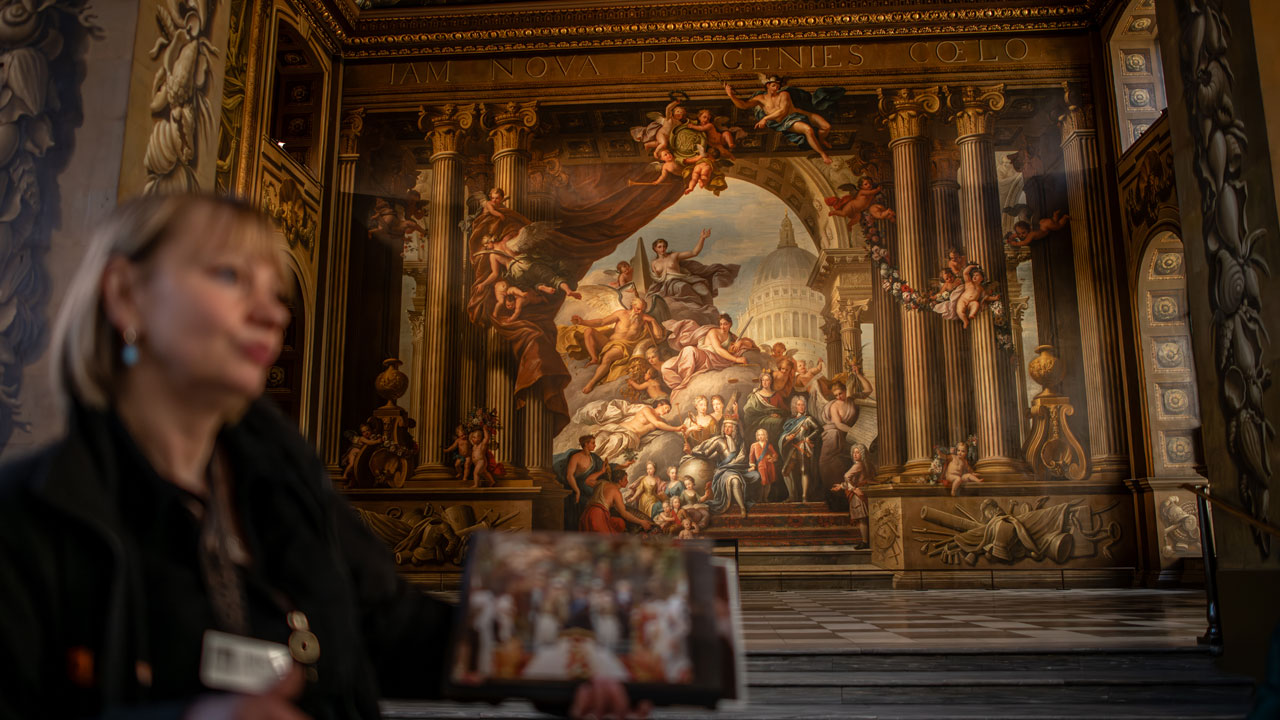 Profitez d'une visite guidée du Painted Hall à Greenwich et découvrez-en plus sur l'envers du décor de ce lieu de tournage emblématique. ©London & Partners/Michael Barrow Une guide conférencière montre une photo d'une scene filmée à l'intérieur du Painted Hall à Greenwich.