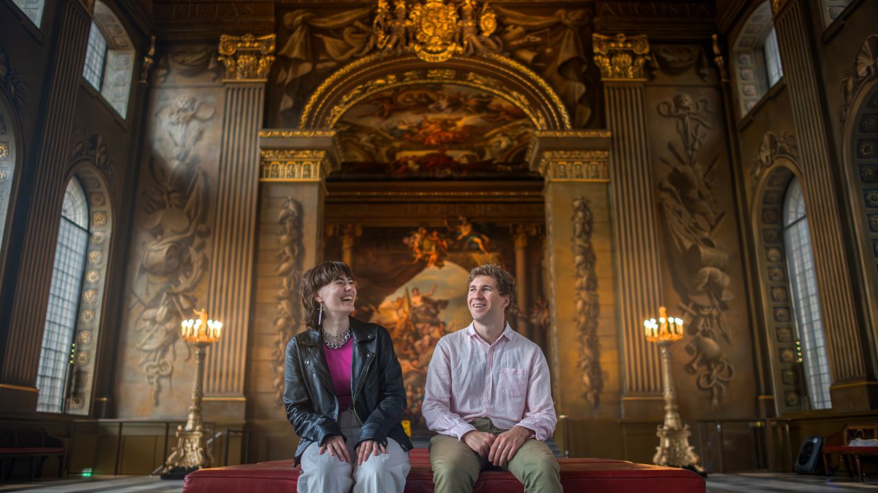 Explorez le Painted Hall, un incontournable lors d'une visite de Greenwich. © London & Partners/Michael Barrow Un homme et une femme sont assis sur un banc et admirent les peintures de style renaissance sur le plafond du Painted Hall à Greenwich.
