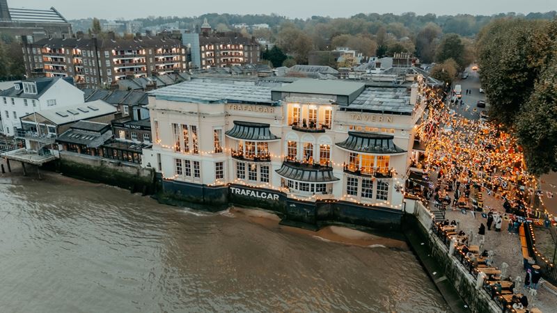 Join the crowds that flock to the vibrant Greenwich riverside to soak up the nightlife at Trafalgar Tavern. Credit: Visit Greenwich. Image courtesy of Visit Greenwich. A bird's-eye view of the Trafalgar Tavern riverside pub in Greenwich with warm fairy lights lining the outdoor area and crowds of people stood and sat by the river Thames