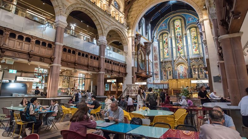 Try incredible street food inside a renovated church at Mercato Mayfair. Image courtesy of Mercato. Diners sit at colourful tables enjoying food inside a renovated church with a high ceiling and stained glass windows at mercato mayfair in london.