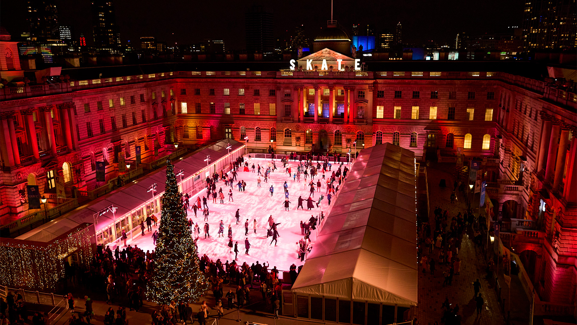 Skate at Somerset House. Image courtesy of Owen Harvey/Somerset House. Skaters glide across the ice at Somerset House against a glittering spruce tree and red lights