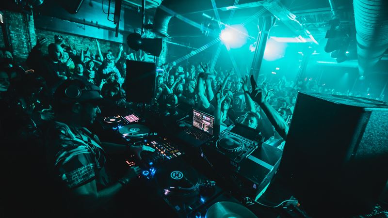 XOYO in Shoreditch is renowned for attracting cutting-edge house, techno and dance acts. Credit: Andy C @Brendon. Image courtesy of XOYO. DJ spinning tunes in front of a crowd dancing with their arms up in the air in a nightclub lit up in green.