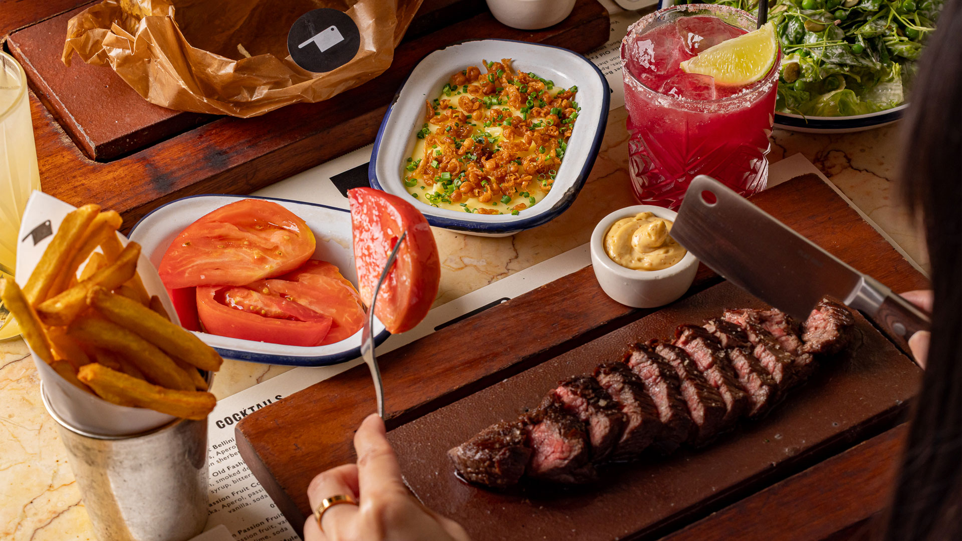Steak and side dishes. Credit: Justin de Souza. Image courtesy of Flat Iron. Sliced steak on a wooden board surrounded by side dishes including chips, tomatoes and macaronic cheese.