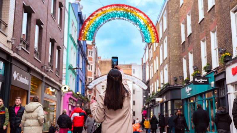Shop until you drop on London's lively Carnaby Street. Photo credit: London & Partners. A woman in a white coat takes a photo of a rainbow decoration on Carnaby street in London