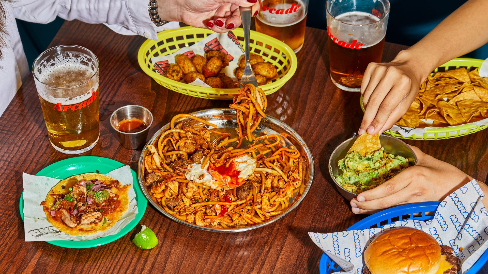 Grab street-food favourites and craft drinks at Arcade Battersea – a lively spot for every craving. Image courtesy of The Cast. A table at Arcade Food Hall with tacos, noodles, guacamole, beers and shared plates.