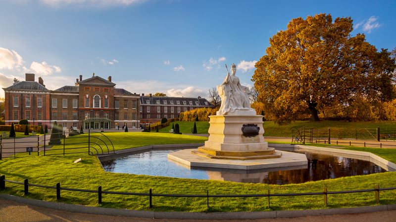 Explore Kensington Gardens in the autumn. Photo: visitlondon.com/Jon Reid. Queen Victoria statue in Kensington Gardens, with tree with autumn leaves, and Kensington Palace in the background