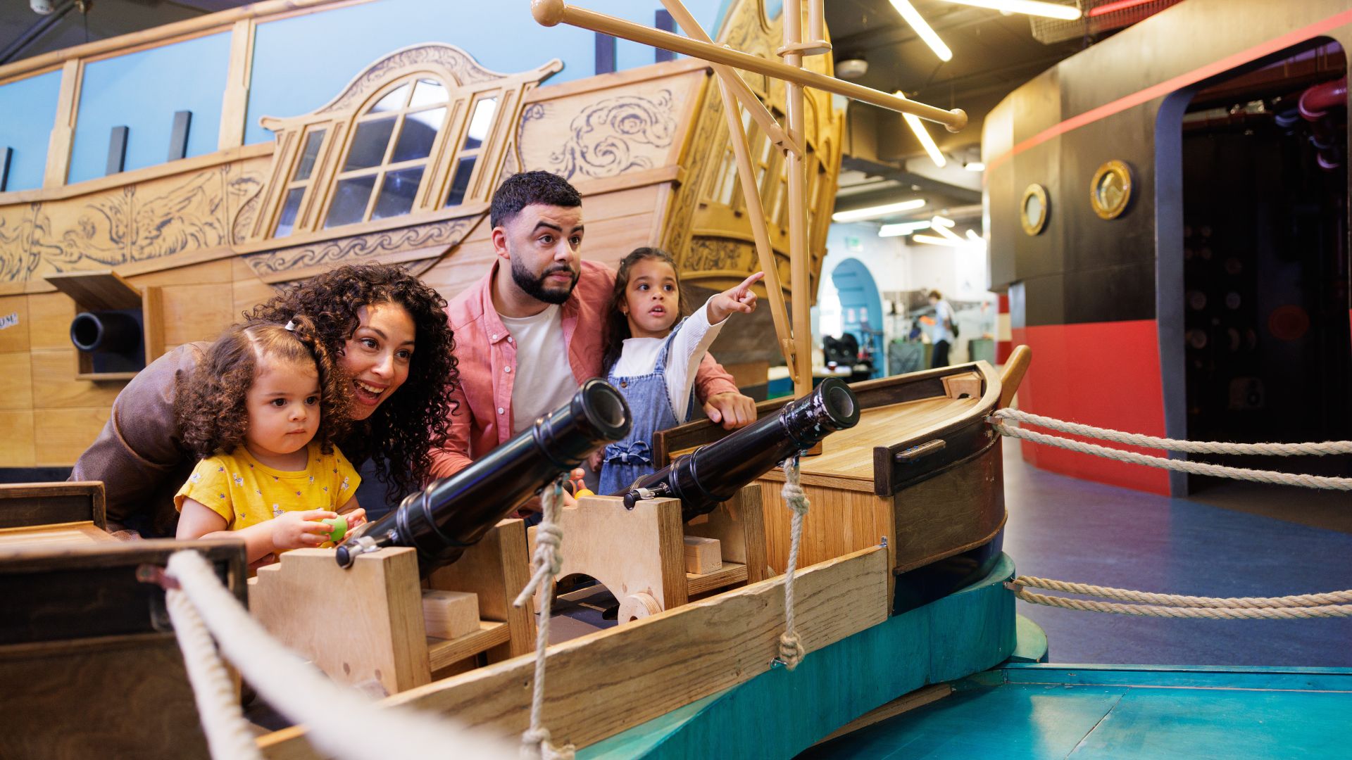 The whole family can have fun at AHOY! at the National Maritime Museum. © National Maritime Museum, Greenwich, London A family of four play at AHOY, firing cannons by a ship, at the national maritime museum in London.