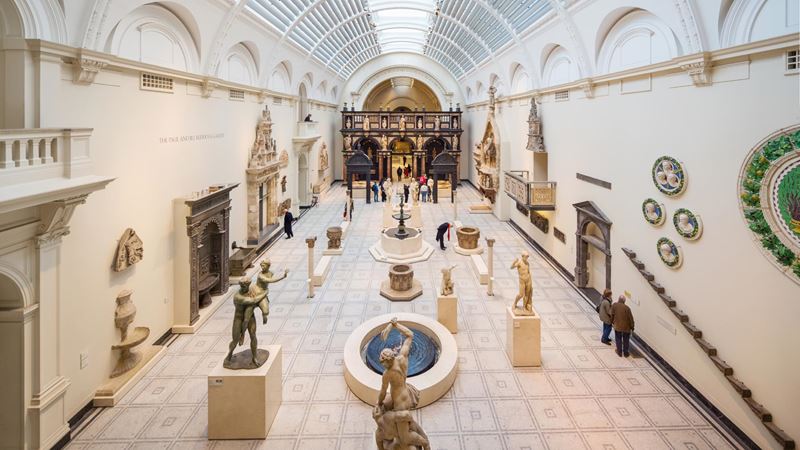 Victoria and Albert Museum. Credit: Jon Reid. Image courtesy of visitlondon.com. A vast light-filled white hall at Victoria and Albert Museum, featuring sculptures and statues on pedestals, artefacts on walls and a curved, glass roof.