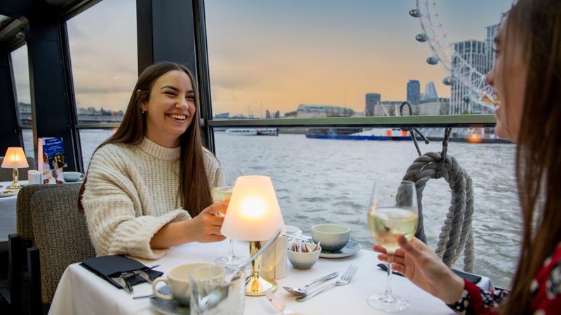 Experience the magic of London at night with a Thames jazz dinner cruise. Credit: Golden Tours. Image courtesy of Golden Tours. two women sat at a table on a cruise boat, cheersing with wine glasses and the sun setting in the background over the London Eye