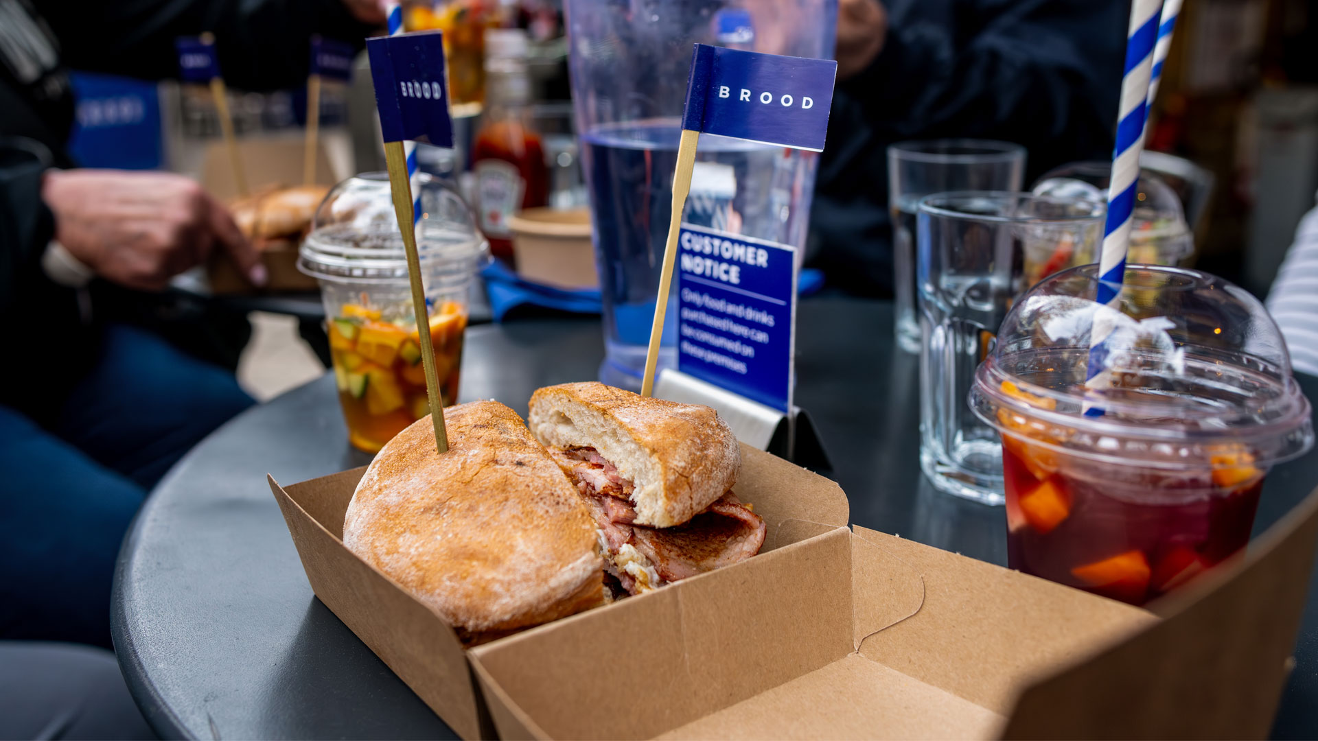 Discover British classics on the Borough Market Food Tour. Bacon sandwich on table surrounded by drinks and people sitting round table at Borough Market.