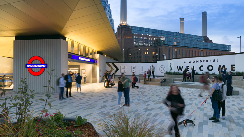 Travel across London using the city's reliable Tube network. Credit: Brendan Bell. Image courtesy of Brendan Bell. Battersea Power Station Tube station in the evening with people walking around in the foreground and the iconic Battersea Power Station with lights on in the background
