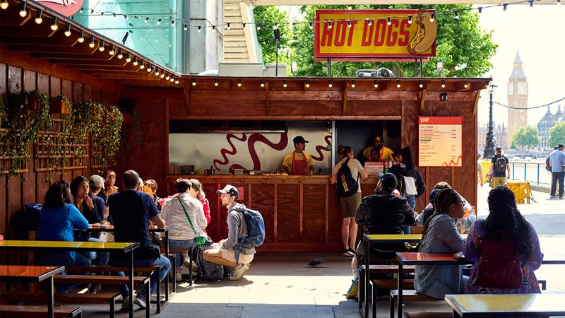 Southbank Centre's riverside food and drink pop-ups serve up foodie delights each summer. Credit: Southbank Centre. Image courtesy of Southbank Centre. A street food stand serving American-style hot dogs with Big Ben pictured in the background.