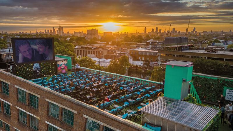 Make London's sunset the backdrop to your next movie date. Credit: Rooftop Cinema Club. Image courtesy of Rooftop Cinema Club. A photo from above Rooftop Film Club in Peckham with the screen playing a movie to seats of people on the rooftop as the sun sets behind London's buildings in the distance