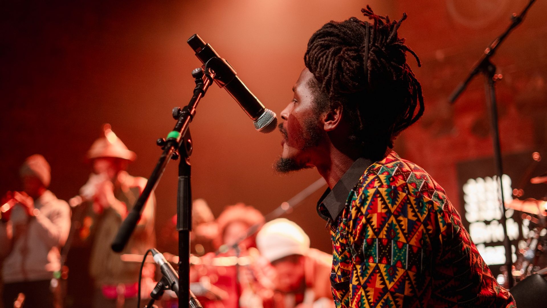 Listen to London's best underground music at Brick Lane Jazz Festival. Credit: Ilhan Beyazay. Image courtesy of Brick Lane Jazz Festival.. A black musician singing live with his band in the background and dim red lighting at the Brick Lane Jazz Festival.