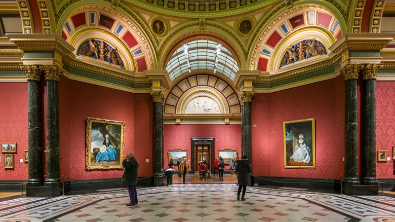 Explore London's National Gallery on a guided tour. Image courtesy of the National Gallery. Two people looking at paintings in the red-painted halls of the National Gallery.