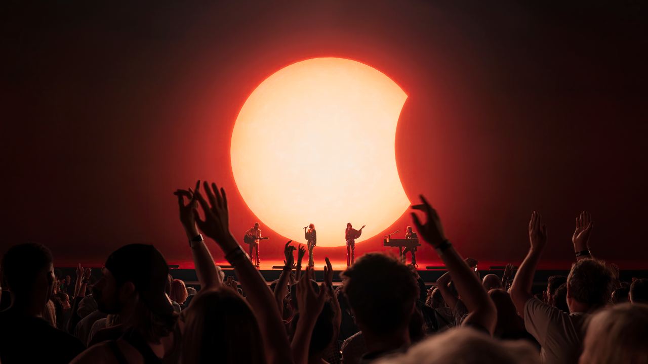 ABBA Voyage performance of Chiquitita at London's ABBA Arena. Image courtesy of Johan Persson/ABBA Voyage. ABBA Voyage performers on stage with red moon backdrop and live crowd with their arms up in the air at London's ABBA Arena.