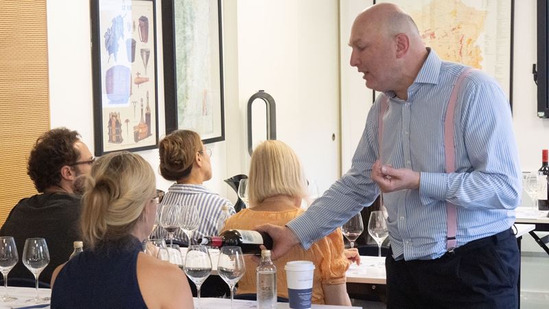 Take part in tastings and workshops as part of Le Cordon Bleu Summer Festival. Image courtesy of Sauce Communications. A man pours a glass of wine for a woman during a wine tasting in a classroom, with various empty glasses on each table.