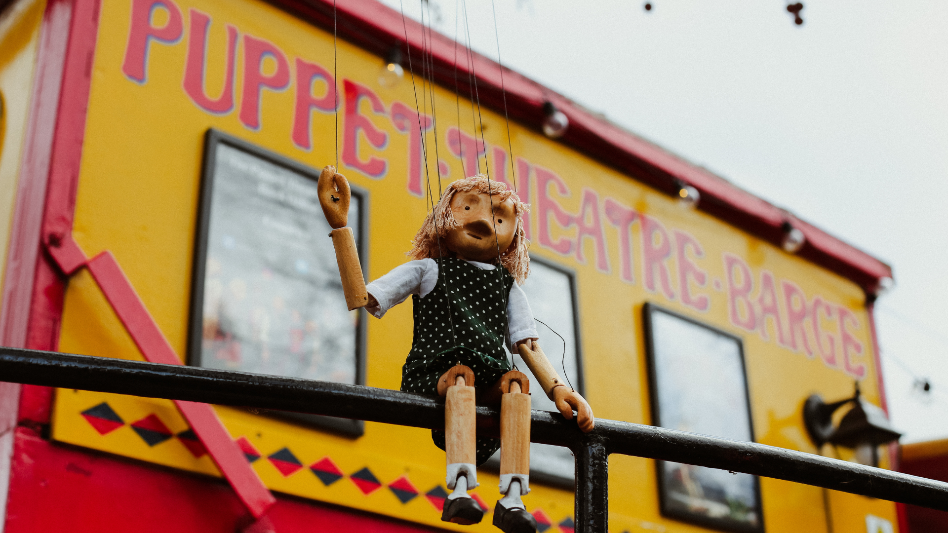 This unique theatre puts on fun shows the whole family can enjoy. Image courtesy of Puppet Theatre Barge. Photo credit: Alex Krook. A blonde puppet sits on a railing in front of the red and yellow puppet theatre barge in London.