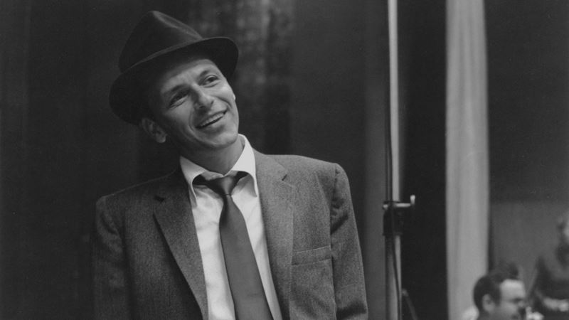 Frank Sinatra pictured during the early years of his career, a defining era explored in Sinatra The Musical. Image courtesy of Story House PR. Black and white photograph of Frank Sinatra wearing a suit and fedora, smiling during a recording session.