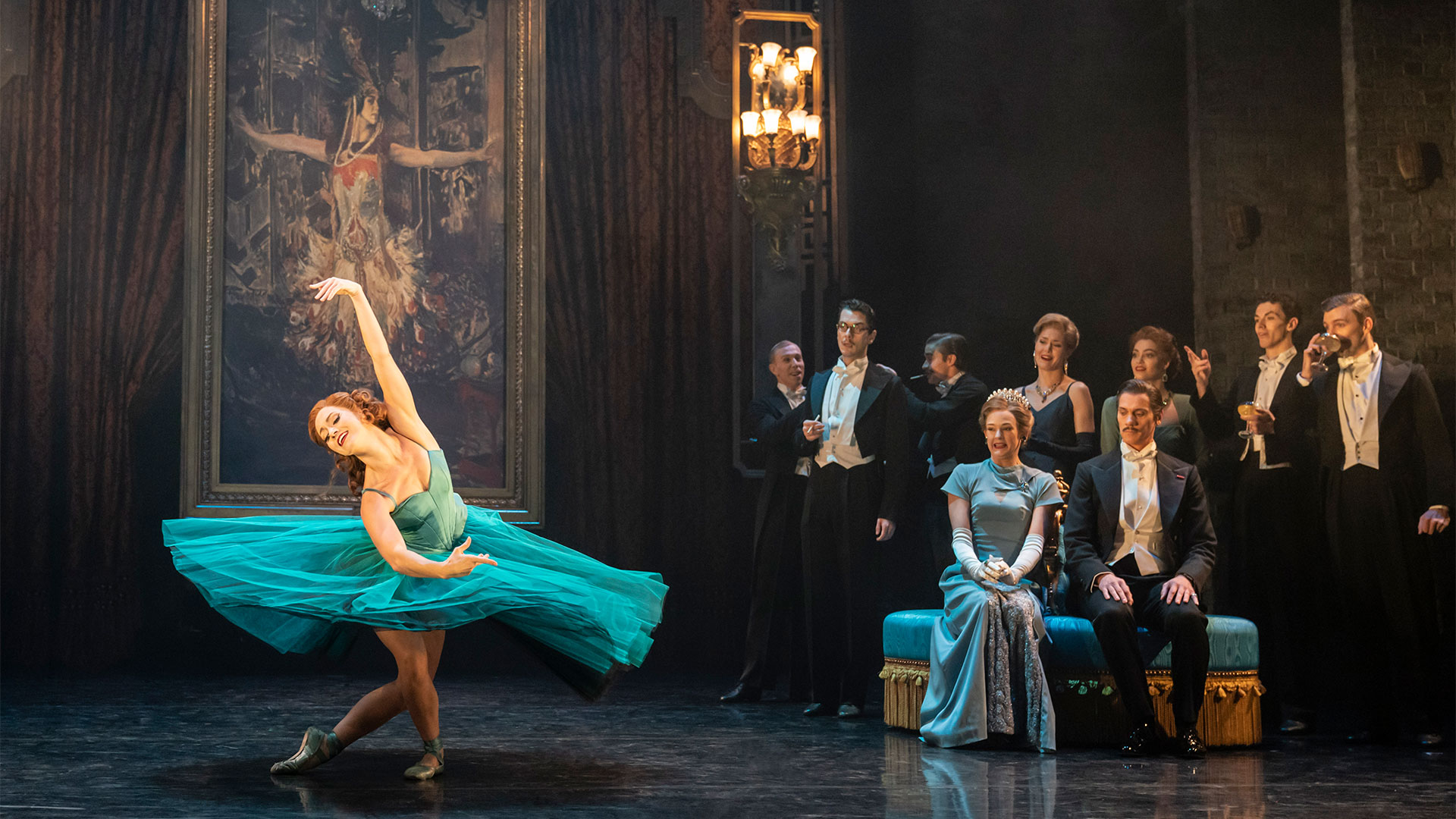 Follow the destiny of a young girl striving to become the best ballet dancer of her generation in Matthew Bourne's new production of The Red Shoes, showing at Sadler's Wells. Photo credit: Johan Persson. A ballerina wearing a bright turquoise tutu dances in front of the other cast members, who are dressed in ball gowns and three-piece suits.