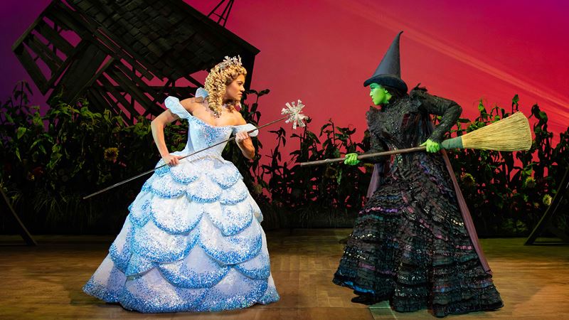 Follow the story of Glinda and Elphaba at Wicked the Musical. Image courtesy of London Theatre Direct/Matt Crockett. Glinda The Good is wearing a light blue gown and is fighting of Elphaba the Wicked Witch who is branding her broom.