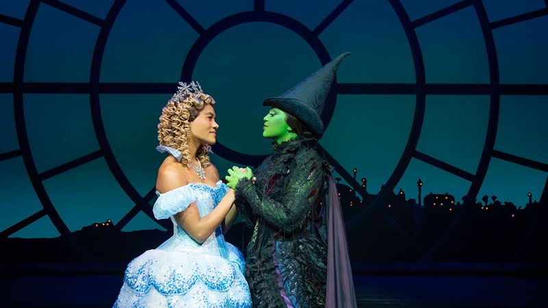 Lucy St Louis et Alexia Khadime interprètent respectivement Glinda et Alphaba dans la comédie musicale de renom Wicked, actuellement à l'affiche à l'Apollo Victoria. Avec l'aimable autorisation de London Theatre DIrect/ crédit photo Matt Crockett. L'actrice qui interprète Glinda dans la comédie musicale Wicked porte une robe de bal bleue et tient la main de l'actrice qui joue le rôle d'Alphaba, célèbre pour sa peau verte, qui porte un chapeau de sorcière et une robe noire.
