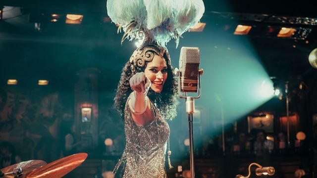A woman dressed in a silver sparkly outfit stands next to a microphone smiling and pointing to the audience.