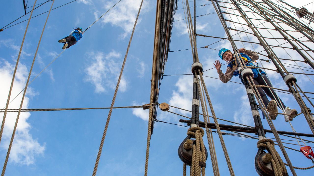 a low angle shot of people wearing jumpsuits and harnesses, climbing the Cutty Sark with a blue sky in the background