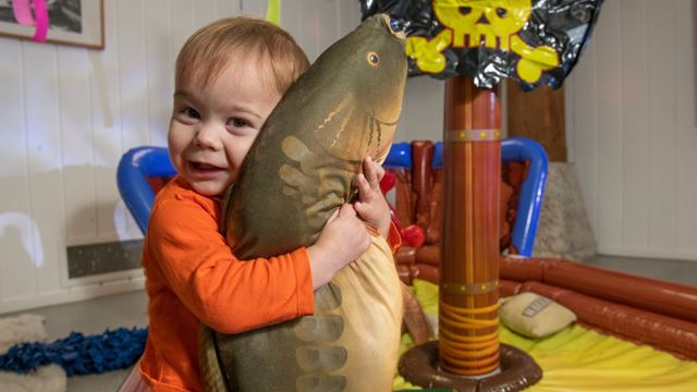 A young child in an orange top hugs a large dinosaur-shaped soft toy inside a colourful indoor play area.