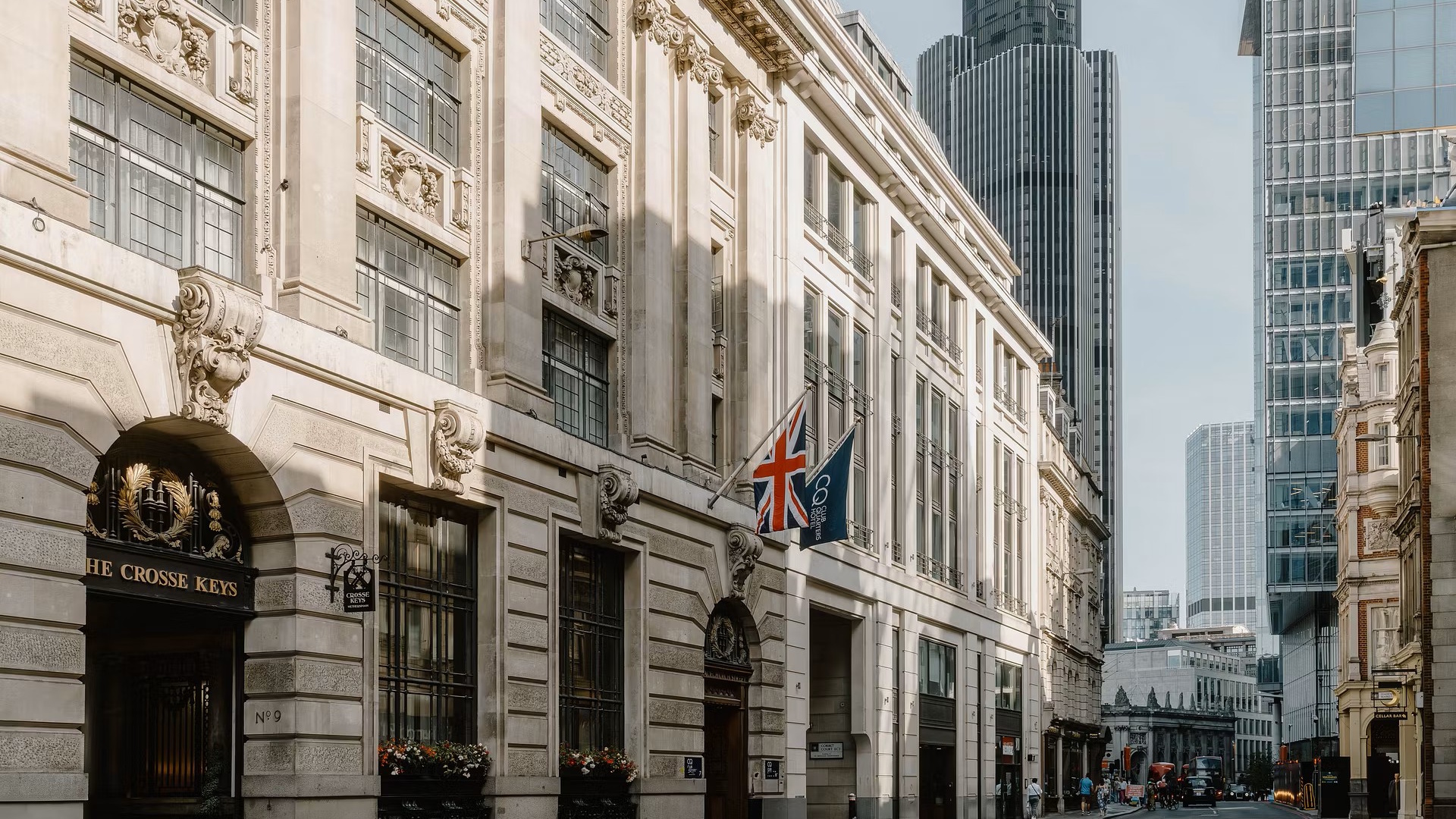 Exterior of Club Quarters hotel in the City of London with the UK and hotel flags above the entrance and skyscrapers and blue sky in the background.