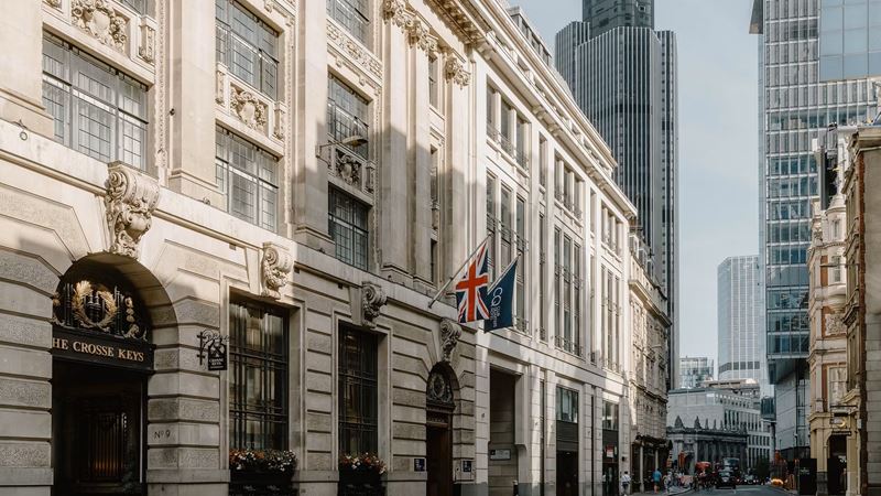 Enjoy a comfy stay at Club Quarters Hotel London St Paul's. Credit: Club Quarters. Image courtesy of Club Quarters. Exterior of Club Quarters hotel in the City of London with the UK and hotel flags above the entrance and skyscrapers and blue sky in the background.