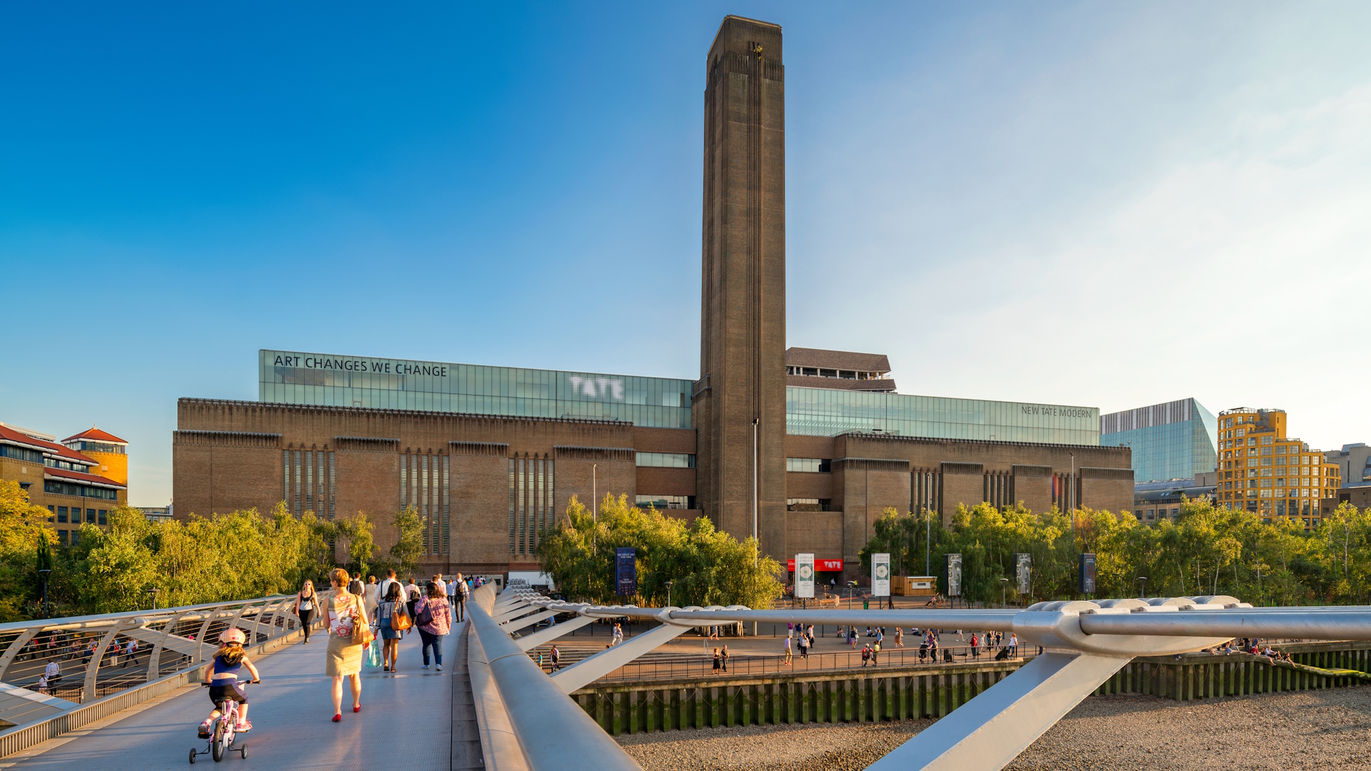 View of Tate Modern building and facade with pedestrians on Millennium Bridge leading up to it, trees and clear blue skies.