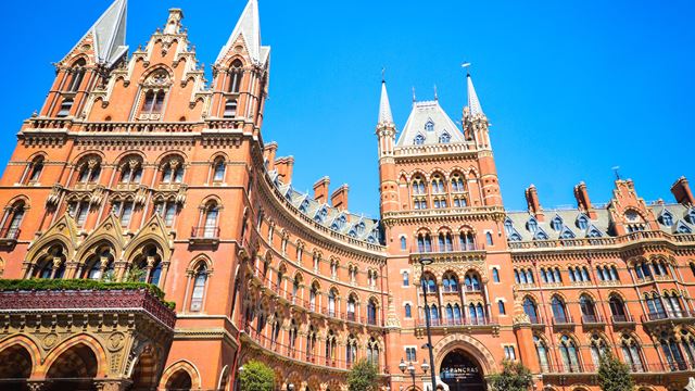 A red-brick, neo-gothic building with two towers, multiple spires and a bright blue sky in the background.