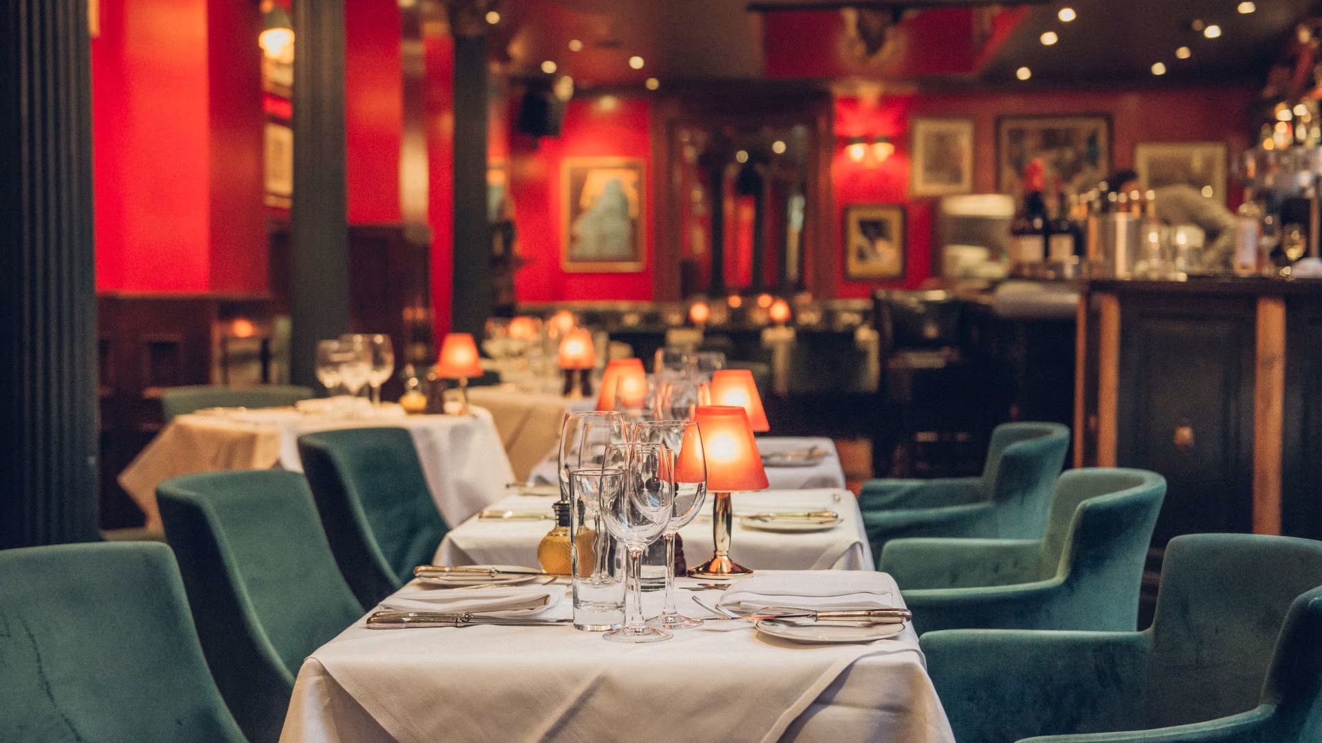 Dining tables lined up with white tablecloth, wine glasses, green velvet seating, cosy lighting and artwork on red walls.