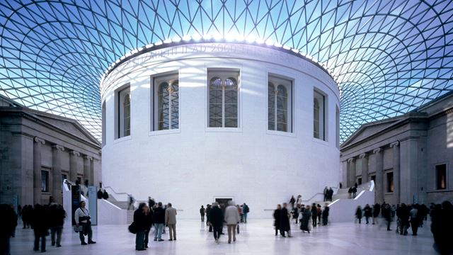 The Great Court at the British Museum with blue sky and light peering through the glass rood, round white structure in the middle, visitors all around and Greek-style buildings in the background.
